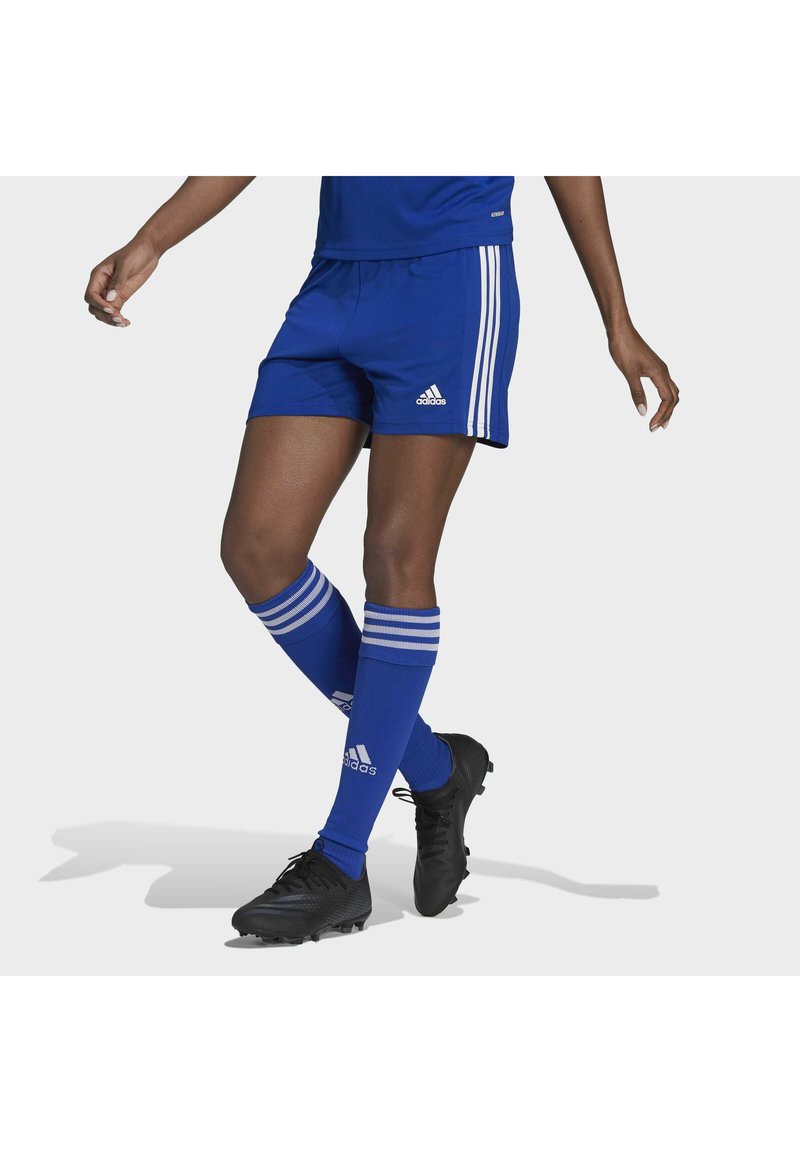 Osoba nosi plave Adidas nogometne šortse, odgovarajuće čarape do koljena i crne kopačke dok odlučno kroči naprijed na jednoličnoj pozadini.
