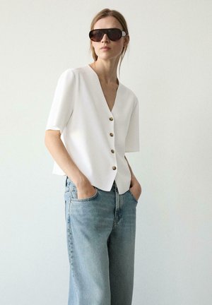 Vrouw met donkere oversized zonnebril, witte blouse met korte mouwen en knopen, en losse lichtblauwe spijkerbroek met handen in de zakken.