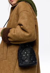 Bolso tipo cubo negro con remaches plateados, hecho de un material suave. El bolso se sostiene contra un abrigo color canela de ante sintético con detalles de pelo sintético verde.