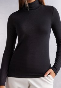 Haut noir à manches longues avec col roulé, en tissu lisse et ajusté ; doté d'un col montant et d'un léger éclat, associé à un pantalon clair.