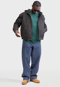 Dark gray zip-up jacket with a hood, worn over a green shirt, paired with loose blue jeans and tan footwear, standing against a plain background.