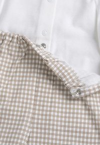 Haut en tissu blanc avec boutons-pression et doublure beige à motif quadrillé, présentant une texture douce et un design ajusté avec une taille élastiquée.