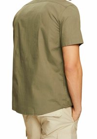 Man in een olijfgroene shirt met korte mouwen en beige cargoshorts, staande met één hand in zijn zak, met de rug naar de kijker.