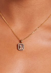 Gouden ketting met een vierkante hanger met een heldere steen in het midden, omringd door kleine sprankelende accenten, tegen een huidskleurige achtergrond.