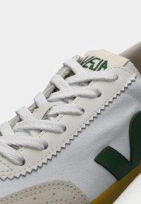 Άσπρο αθλητικό παπούτσι με πράσινο λογότυπο, επάνω μέρος από καμβά, λευκά κορδόνια, υφή εξωτερικής σόλας από καουτσούκ και κρεμ σουέντ λεπτομέρειες.