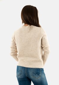 Une femme aux longs cheveux bruns porte un pull beige et un jean bleu, de dos contre un fond blanc uni.