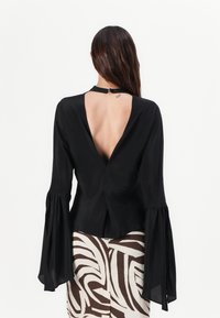 Black blouse with a deep V-back, long flared sleeves, and a button closure at the neck. Features smooth, lightweight fabric.