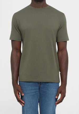 Mand iført en ensfarvet olivengrøn T-shirt med kortærmet rund hals og blå jeans, stående mod en neutral baggrund.