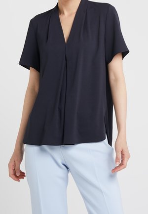 Mujer con blusa holgada de manga corta y cuello en V de color azul marino oscuro, combinada con pantalones azul claro de corte entallado, sobre un fondo liso.