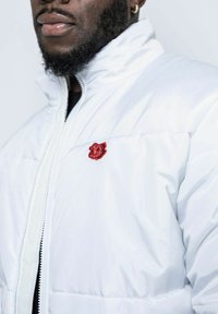 Vit vadderad jacka med hög krage. Har en röd broderad ros på vänster sida. Slät textur, dragkedja och en något puffig design.