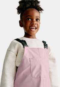 Salopette imperméable rose avec poche frontale, dotée de bretelles noires ajustables. Portée par-dessus une chemise à manches longues légère et tachetée.