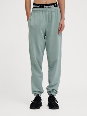 Osoba ubrana w jasnozielone spodnie dresowe i czarne tenisówki, stojąca ze zrelaksowanymi rękami wzdłuż ciała na białym tle.