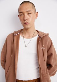 Sudadera con cremallera marrón y detalles en blanco, llevada sobre una camiseta de tirantes blanca con textura acanalada. Collares de cadena plateada. Textura de tela suave con bolsillo tipo canguro.