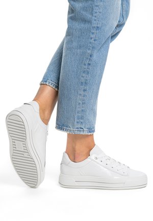 Jambes en jean bleu clair portant des baskets basses blanches, un pied à plat et l'autre levé montrant la semelle de la chaussure avec un motif rainuré.