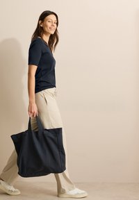 Duża czarna torba na zakupy z dwoma uchwytami. Gładka tekstura, minimalistyczny design. Model ma na sobie granatową koszulę i jasnobeżowe spodnie.