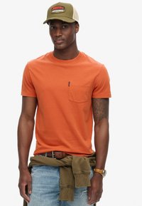 Orange bomulds T-shirt med rund hals og lomme, båret med afslappede lyseblå jeans. En khaki kasket med et logo fuldender looket.