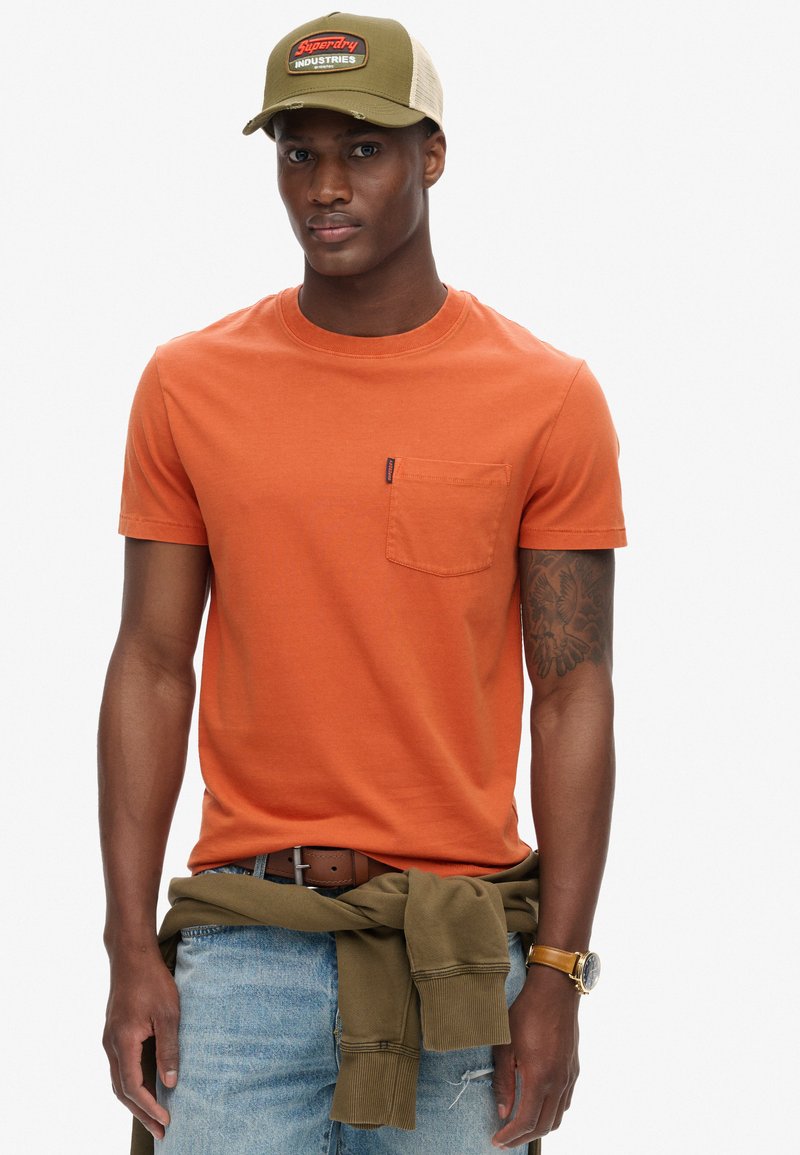 Orange bomulds T-shirt med rund hals og lomme, båret med afslappede lyseblå jeans. En khaki kasket med et logo fuldender looket.