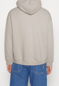 Felpa grigia chiara con cappuccio regolabile, vestibilità comoda e polsini a costine. Il tessuto appare morbido e strutturato, abbinato a pantaloni in denim blu.