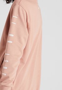Sweatshirt rose à manches longues en tissu doux. Il présente le texte blanc "JUST DO IT." le long de la manche gauche et un petit logo blanc sur la poitrine.