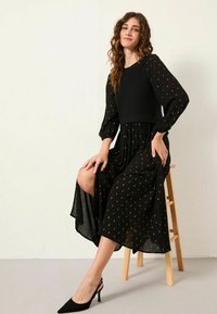 Schwarzes Kleid mit langen, gepufften Ärmeln und goldenen Mustern. Durchsichtiger unterer Teil mit seitlichem Schlitz. Kombination mit schwarzen High Heels.