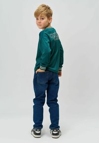 Cămașă din teal cu mânecă lungă, guler în dungi și logo pe spate, asortată cu blugi de culoare albastru închis și adidași negri, purtată de un copil pe un fundal simplu.