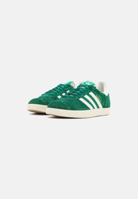 Green suede sneakers with white leather accents, featuring three white stripes, a round toe, and a textured rubber sole. "Gazelle" embossed on side.
