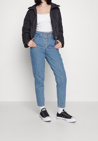 Veste rembourrée noire, crop top blanc froncé, jeans taille haute bleus avec fermeture à boutons, et baskets en toile noires avec semelles blanches.