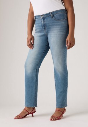 Kvinde iført lyseblå jeans med lige ben og røde sandaler med lave hæle og stropper, stående foran en ensfarvet baggrund.