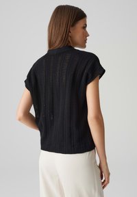 Top in maglia nera con maniche corte, caratterizzato da un motivo testurizzato e un design a trama aperta. Il retro è leggermente più lungo della parte anteriore.