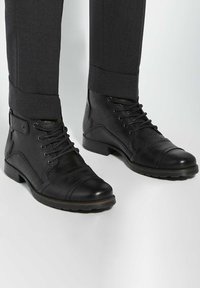 Black leather ankle boots with a smooth finish, featuring laces, a cap toe, and a rubber sole. Worn with charcoal grey tailored trousers.