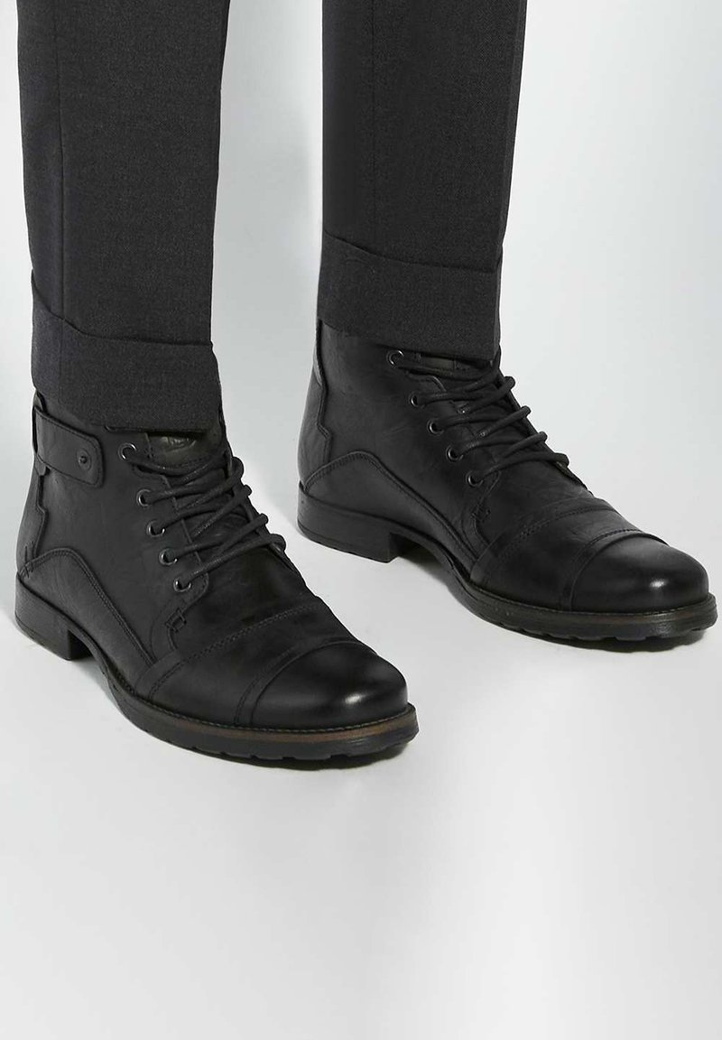 Black leather ankle boots with a smooth finish, featuring laces, a cap toe, and a rubber sole. Worn with charcoal grey tailored trousers.