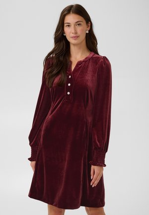 Jeune femme portant une robe en velours bordeaux à manches longues avec des boutons décoratifs et des poignets smockés, debout devant un fond uni.