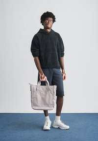 Νεαρό άτομο με λεύκη που φοράει μαύρη κουκούλα και σορτς, κρατώντας μπεζ τσάντα tote, στέκεται σε μπλε χαλί με φόντο λευκό τοίχο.