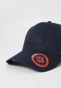 Dunkelblaues Baseballcap mit gesticktem "HUGO"-Logo. Verfügt über einen gebogenen Schirm und ein runder Anhänger mit "HUGO"-Branding in Orange und Schwarz.