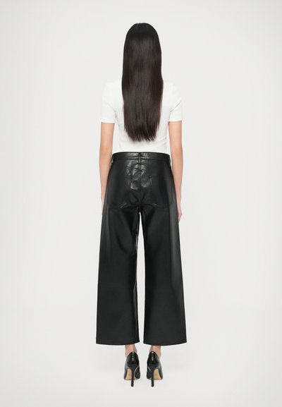 Schwarze Lederhose mit weitem Bein und hoher Taille, ausgestattet mit zwei Gesäßtaschen und einer glatten Oberfläche. Kombiniert mit schwarzen Schuhen mit Absatz.