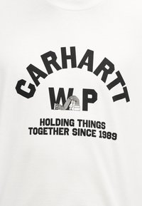 Λογότυπο Carhartt WIP με ένα άτομο να κοιτάζει ανάμεσα στα γράμματα και κείμενο «Κρατώντας τα πράγματα μαζί από το 1989» σε λευκό ύφασμα.