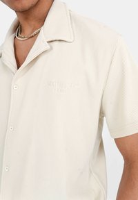Camisa beige de manga corta con textura acanalada, cuello con muesca y logotipo bordado en el pecho. Los botones blancos acentúan el frente.