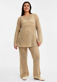 Beige meshstickad tvådelad set bestående av långärmad topp och utsvängda byxor med lös passform. Tyget har ett öppet, texturerat mönster.