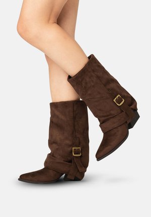 Ben iført mellemlange mørkebrun ruskindsslouchy-støvler med en sidelås og messing spænde, lave stablede hæle, spidse tæer.