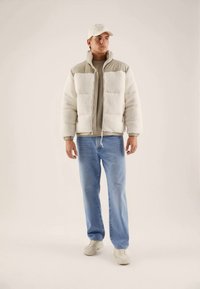 Gevoerde crèmekleurige fleecejas met beige accenten, gecombineerd met een beige shirt en blauwe jeans. Model draagt een lichte pet en beige sneakers.