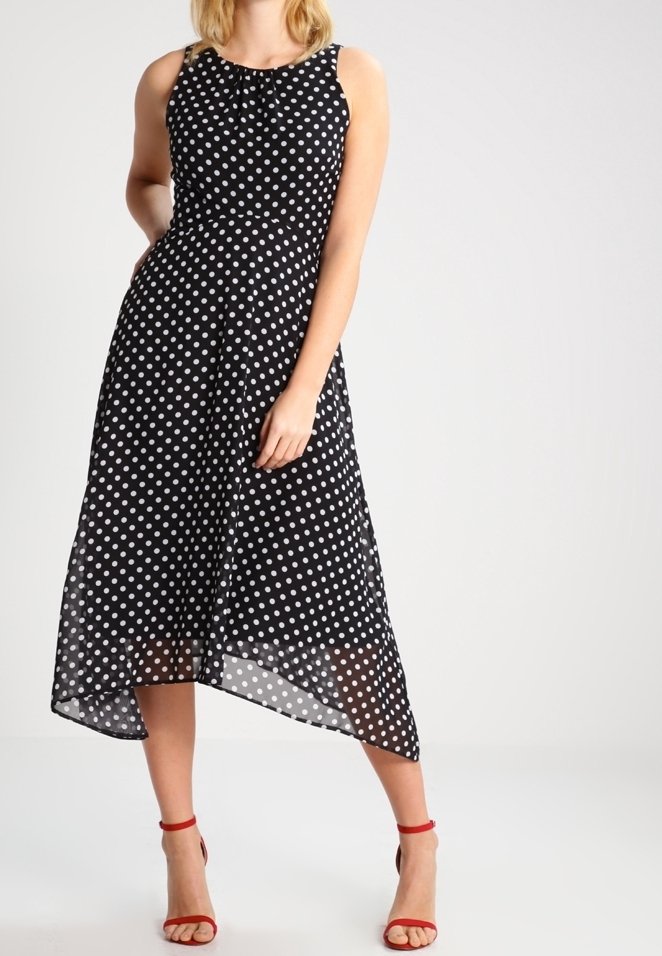 Femme en robe noire sans manches à pois blancs, ourlet asymétrique, avec des sandales rouges à talons hauts, debout devant un fond uni.