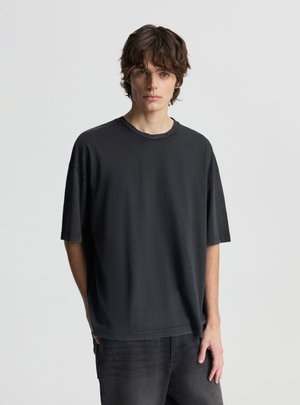 Młody mężczyzna z falującymi brązowymi włosami, ubrany w luźną czarną koszulkę i ciemne dżinsy, stojący na tle jednolitego jasnoszarego tła.