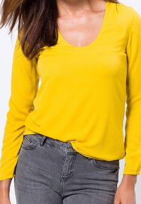 Gul bluse med lange ærmer og rund halsudskæring, lavet af et glat materiale. Kombineret med tætsiddende grå jeans med knaplukning.