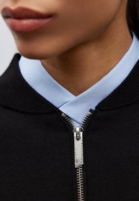 Svart zip-up plagg med ljusblå krage, prydt med en silverdragkedja med ingraverad detalj, samt en strukturerad tyg.