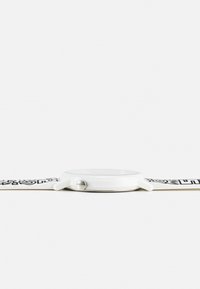 Montre-bracelet blanche avec un boîtier rond fin, un verre plat, une couronne argentée et un bracelet à motif noir et blanc, posée à plat sur une surface blanche.