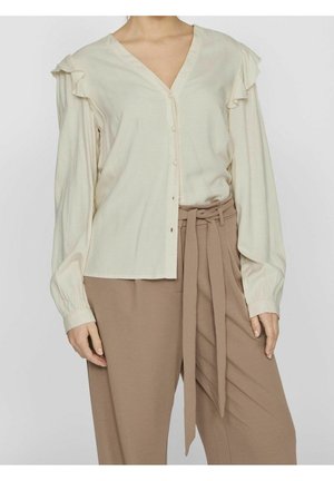 Kobieta ubrana w kremową, zapinaną na guziki bluzkę z falbankami na ramionach oraz brązowe spodnie z wysokim stanem i przewiązanym paskiem z tkaniny.