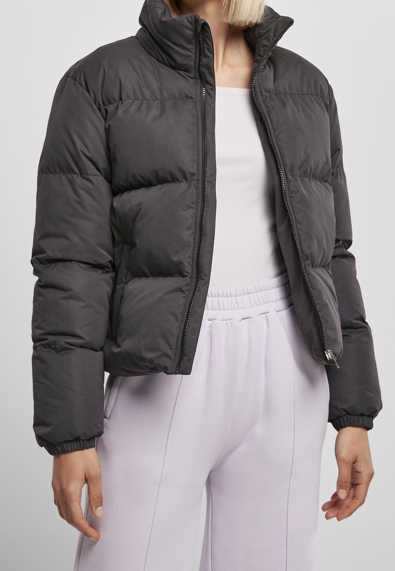 Femme portant une doudoune noire par-dessus un haut blanc et un pantalon de survêtement violet clair avec une ceinture élastique, debout devant un fond uni.