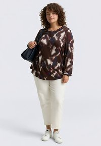 Maglia marrone a maniche lunghe con motivo astratto bianco e blu, abbinata a pantaloni bianchi e sneakers bianche, con una borsa nera a tracolla.