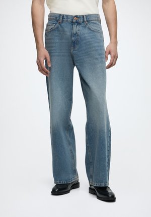 Personne portant un jean bleu amples avec des zones décolorées et des chaussures en cuir noires, debout devant un fond clair uni.