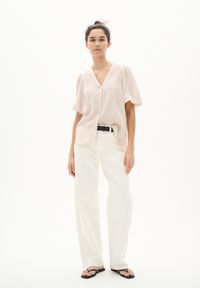 Blusa rosa chiaro a maniche corte con scollatura a V e chiusura a bottone, abbinata a pantaloni bianchi a gamba larga e ciabatte nere su uno sfondo neutro.